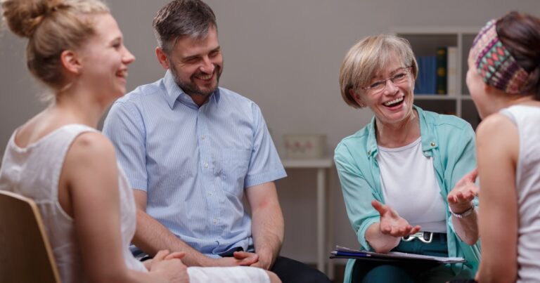 Diverse adults laughing together in group therapy session with female therapist facilitating discussion in modern treatment facility