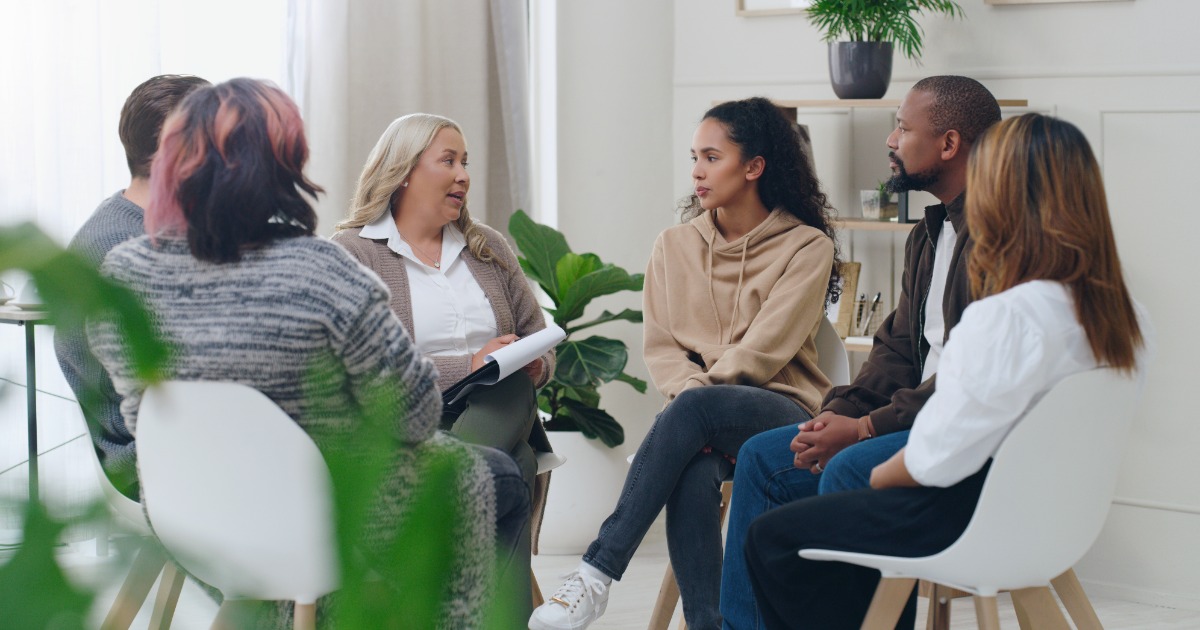 Diverse group therapy session for addiction recovery with counselor and participants in comfortable modern setting with natural lighting and plants