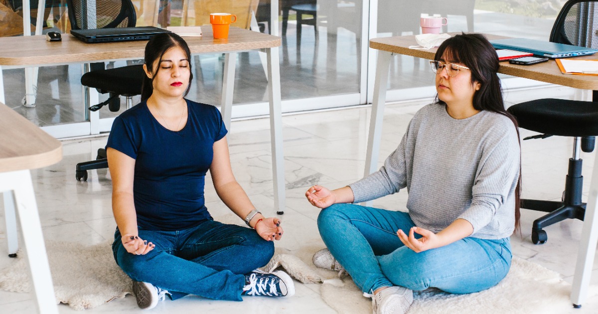 Two women practicing breathing exercises together for anxiety support, sitting on floor in comfortable workspace