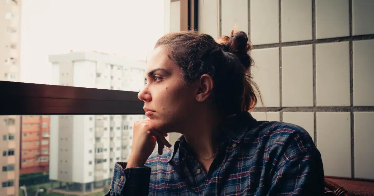 Young woman sitting thoughtfully by window contemplating early signs of bipolar disorder and mental health concerns